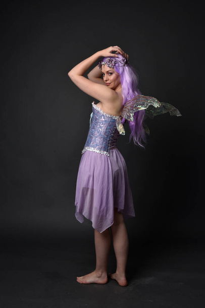 Täyspitkä muotokuva violetti tukka tyttö yllään fantasia korsetti mekko keiju siivet ja kukka kruunu. Seisovat vasten tumma studio tausta. - Valokuva, kuva