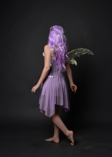 Retrato de larga duración de una niña de pelo morado con vestido de corsé de fantasía con alas de hadas y corona de flores. Posar de pie sobre un oscuro fondo de estudio. - Foto, imagen