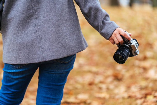 close up female traveller hand hold camera in one hand standing in autumn brown falling tree leaf public nature garden park travel concept - Foto, afbeelding