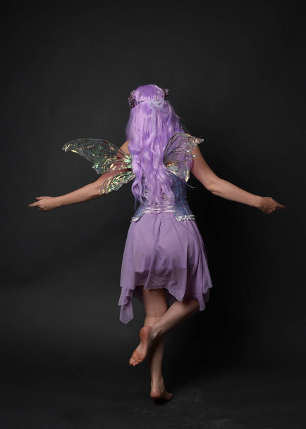 Retrato de larga duración de una niña de pelo morado con vestido de corsé de fantasía con alas de hadas y corona de flores. Posar de pie sobre un oscuro fondo de estudio. - Foto, Imagen