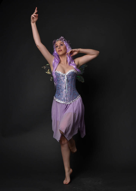 Retrato de comprimento total de uma menina de cabelos roxos vestindo vestido de espartilho fantasia com asas de fadas e coroa de flores. pose de pé contra um fundo de estúdio escuro. - Foto, Imagem