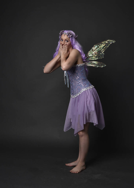 Ganztägiges Porträt eines lila haarigen Mädchens in einem fantasievollen Korsettkleid mit Feenflügeln und Blumenkrone. Stehende Pose vor dunklem Studiohintergrund. - Foto, Bild