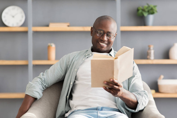 Μείνετε σπίτι χόμπι. Χαρούμενος αφροαμερικανός ώριμος άντρας που απολαμβάνει το διάβασμα βιβλίου, κάθεται σε αναπαυτική πολυθρόνα, ελεύθερος χώρος - Φωτογραφία, εικόνα