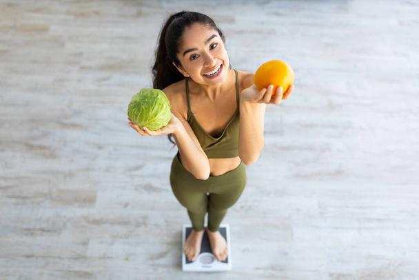 Healthy diet for weight loss. Above view of millennial Indian lady holding citrus fruit and vegetable while using scales - Photo, Image