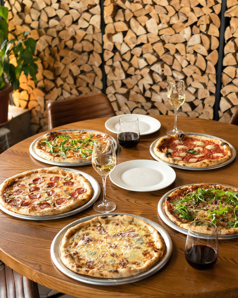 Festa das pizzas. Sorteado cinco pizzas diferentes em mesa de restaurante de madeira com copos de vinho sobre ele. Pasta de lenha sobre fundo desfocado. Modelo para banner de pizzaria. Formato vertical - Foto, Imagem