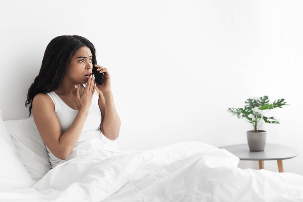 Geschokte zwarte vrouw zitten op bed en praten op de smartphone, op zoek naar kopieerruimte, slaapkamer interieur - Foto, afbeelding