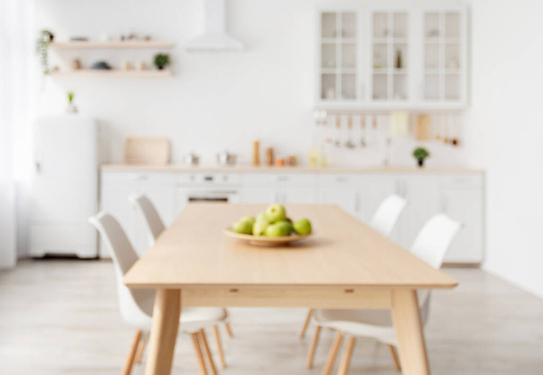 Θολή φόντο με σύγχρονο φως κουζίνα, ξύλινο τραπέζι και λευκά έπιπλα, καρέκλες, μαγειρικά σκεύη και σκεύη - Φωτογραφία, εικόνα