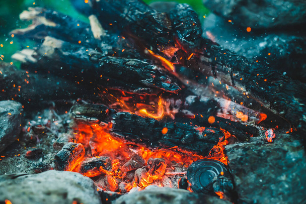 Levendige smeulende brandhout verbrand in brand close-up. Sfeervolle achtergrond met oranje vlam van kampvuur. Volledig beeld van kampvuur met vonken in bokeh. Warme vortex van gloeiende vlammen en as in de lucht - Foto, afbeelding