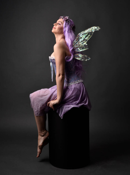Ολόσωμο πορτραίτο ενός μωβ κοριτσιού με φανταστικό κορσέ φόρεμα με φτερά νεράιδας και στεφάνι λουλουδιού. Καθισμένη ποζάρουν σε σκοτεινό φόντο στούντιο. - Φωτογραφία, εικόνα
