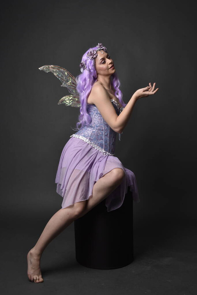 Portrait complet d'une fille aux cheveux violets portant une robe corset fantaisie avec des ailes de fée et une couronne de fleurs. Pose assise sur un fond de studio sombre. - Photo, image