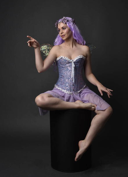 Full length portrait of a purple haired girl wearing fantasy corset dress with fairy wings and flower crown.  Seated pose against a dark studio background. - Photo, Image
