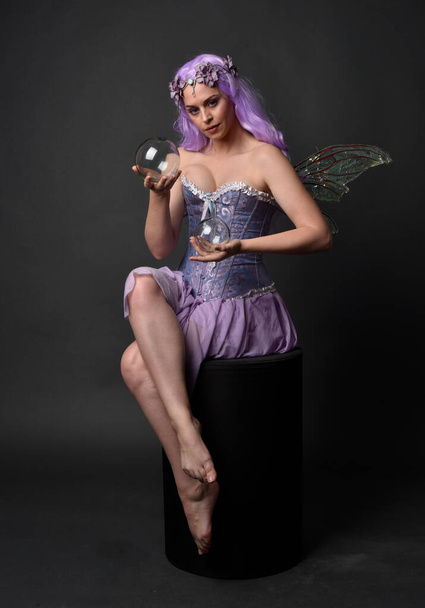 Ganztägiges Porträt eines lila haarigen Mädchens in einem fantasievollen Korsettkleid mit Feenflügeln und Blumenkrone. Sitzende Pose vor dunklem Studiohintergrund. - Foto, Bild