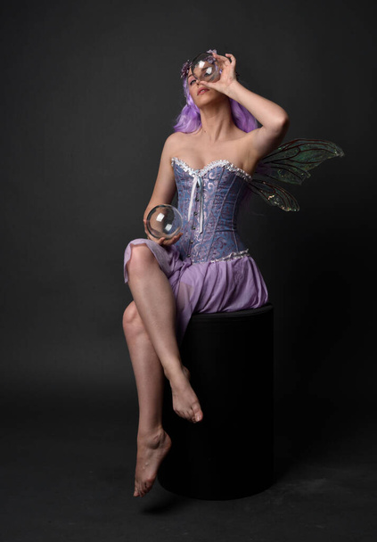Ολόσωμο πορτραίτο ενός μωβ κοριτσιού με φανταστικό κορσέ φόρεμα με φτερά νεράιδας και στεφάνι λουλουδιού. Καθισμένη ποζάρουν σε σκοτεινό φόντο στούντιο. - Φωτογραφία, εικόνα