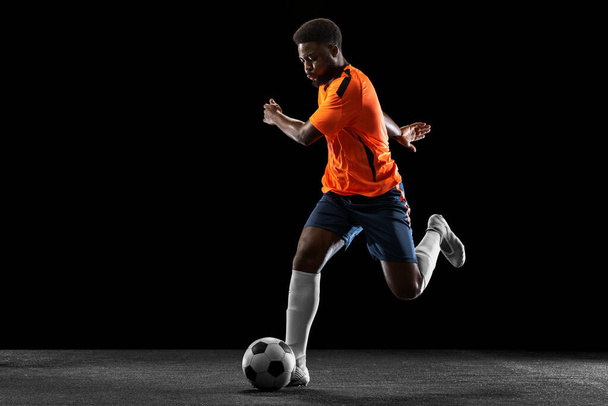 Young African football soccer player playing isolated on black background. Concept of sport, movement, energy and dynamic. - Zdjęcie, obraz