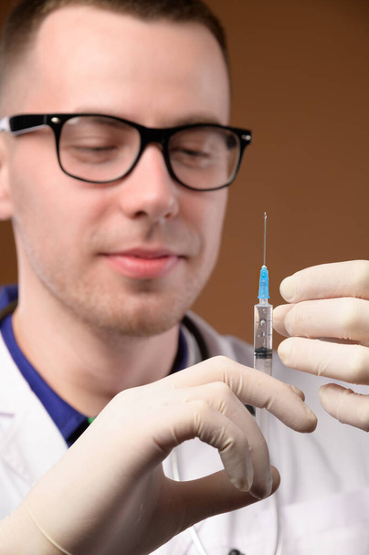 Um jovem médico de óculos com um estetoscópio ao redor do pescoço e com um casaco branco segura uma seringa de plástico com uma injeção. Foco na seringa - Foto, Imagem