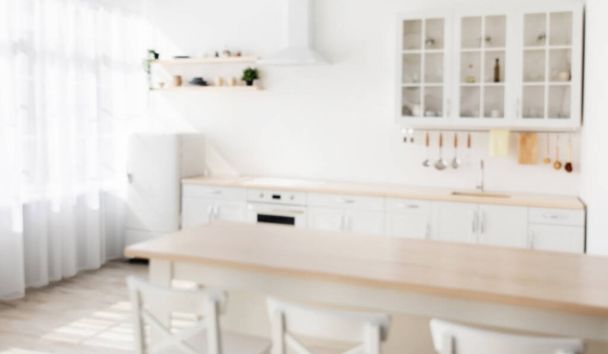 Rozostřené fotografie dřevěného jídelního stolu a bílých židlí v blízkosti, různé nádobí na lehkém kuchyňském nábytku - Fotografie, Obrázek
