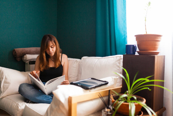 Młoda rudowłosa kobieta, czytająca książkę na kanapie otoczona roślinami. Rozrywka w pomieszczeniach rekreacyjnych. - Zdjęcie, obraz