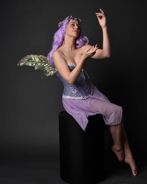 Teljes hosszúságú portré egy lila hajú lányról, aki fantázia fűzőt visel tündér szárnyakkal és virágkoronával. Ült pózban szemben sötét stúdió háttér. - Fotó, kép