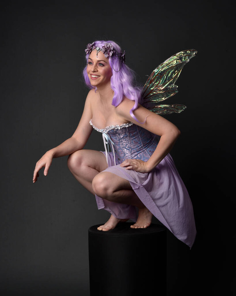 Full length portret van een paars harig meisje dragen fantasie korset jurk met fee vleugels en bloem kroon. Zittende pose tegen een donkere studio achtergrond. - Foto, afbeelding