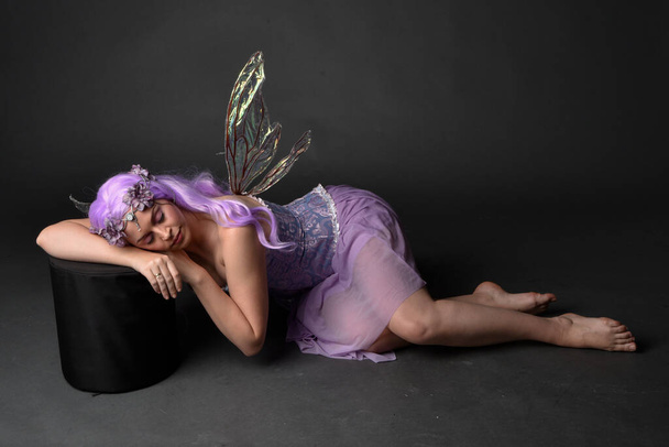 Ganztägiges Porträt eines lila haarigen Mädchens in einem fantasievollen Korsettkleid mit Feenflügeln und Blumenkrone. Sitzende Pose vor dunklem Studiohintergrund. - Foto, Bild