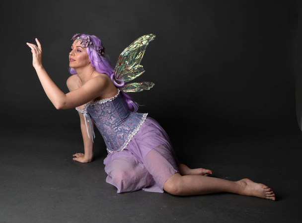 Plně dlouhý portrét fialovlasé dívky ve fantazijních korzetových šatech s vílými křídly a květinovou korunou. Sedící póza na pozadí tmavého studia. - Fotografie, Obrázek
