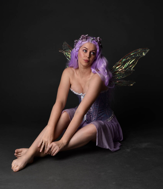 Teljes hosszúságú portré egy lila hajú lányról, aki fantázia fűzőt visel tündér szárnyakkal és virágkoronával. Ült pózban szemben sötét stúdió háttér. - Fotó, kép