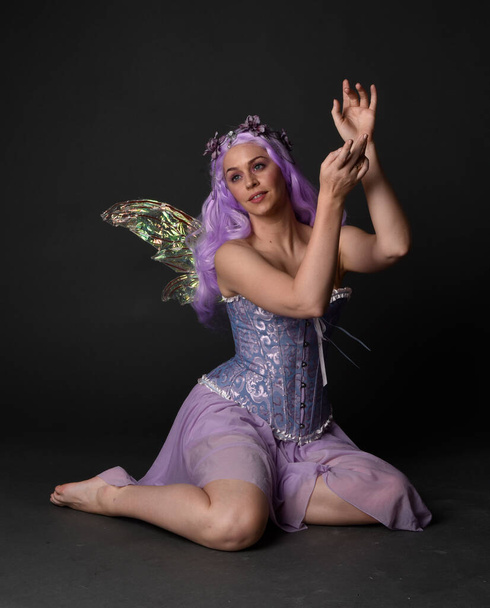 Ritratto a figura intera di una ragazza dai capelli viola che indossa un abito in corsetto fantasia con ali di fata e corona di fiori. Posa seduta sullo sfondo di uno studio buio. - Foto, immagini