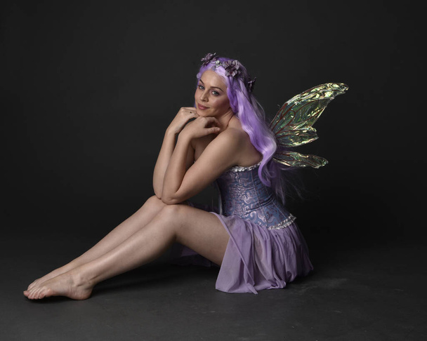 Портрет девушки с фиолетовыми волосами в сказочном корсетном платье с крыльями феи и цветочной короной. Сидячая поза на темном фоне студии. - Фото, изображение
