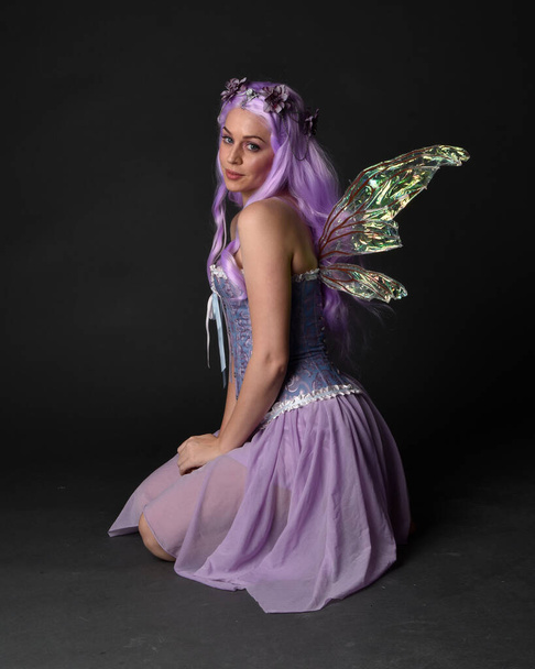 Full length portrait of a purple haired girl wearing fantasy corset dress with fairy wings and flower crown.  Seated pose against a dark studio background. - Фото, изображение