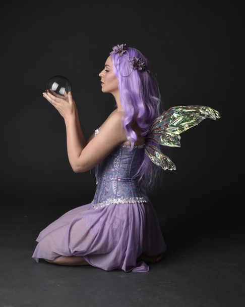Täyspitkä muotokuva violetti tukka tyttö yllään fantasia korsetti mekko keiju siivet ja kukka kruunu. Istuva aiheuttaa vastaan tumma studio tausta. - Valokuva, kuva