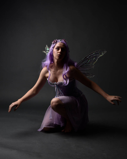 Full length portrait of a purple haired  girl wearing fantasy corset dress with fairy wings and flower crown.  Seated pose against a dark studio background. - Fotoğraf, Görsel