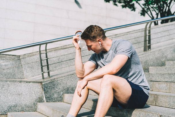Unavený mladý muž běžec sedí a relaxaci po sportovním tréninku. Držení láhve s vodou při cvičení v letní City Street. - Fotografie, Obrázek