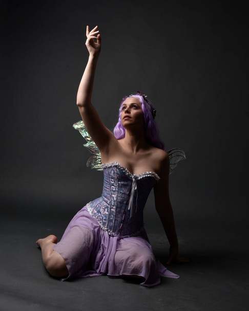Täyspitkä muotokuva violetti tukka tyttö yllään fantasia korsetti mekko keiju siivet ja kukka kruunu. Istuva aiheuttaa vastaan tumma studio tausta. - Valokuva, kuva