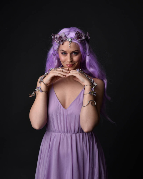 Ritratto ravvicinato di una ragazza dai capelli viola che indossa un abito in corsetto fantasia con ali di fata e corona di fiori, lanciando un incantesimo. In posa su uno sfondo oscuro da studio. - Foto, immagini