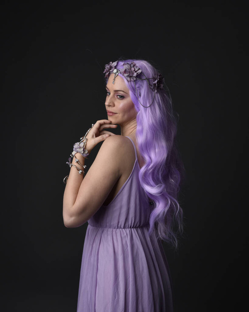 Κοντινό πορτραίτο ενός μοβ κοριτσιού με φανταστικό κορσέ φόρεμα με φτερά νεράιδας και στεφάνι λουλουδιών, που κάνει μάγια. Τοποθέτηση σε σκοτεινό φόντο στούντιο. - Φωτογραφία, εικόνα