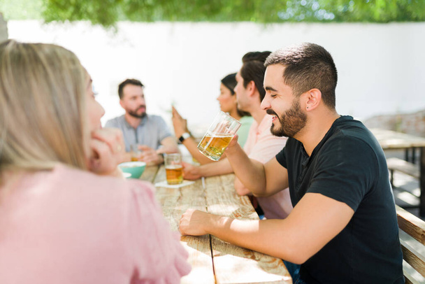 Bromas y risas con amigos. Hombre hispano guapo bebiendo una cerveza fría y relajándose con un gran grupo de amigos en un bar - Foto, Imagen