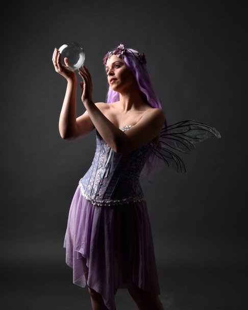 Κοντινό πορτραίτο ενός μοβ κοριτσιού με φανταστικό κορσέ φόρεμα με φτερά νεράιδας και στεφάνι λουλουδιών, που κάνει μάγια. Τοποθέτηση σε σκοτεινό φόντο στούντιο με σκιασμένο φωτισμό backlight. - Φωτογραφία, εικόνα