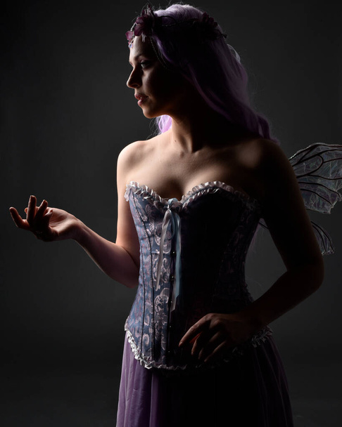 Lähikuva muotokuva violetti tukka tyttö yllään fantasia korsetti mekko keiju siivet ja kukka kruunu, valu loitsu. Poseeraus tumma studio tausta varjoon taustavalaistu valaistus. - Valokuva, kuva