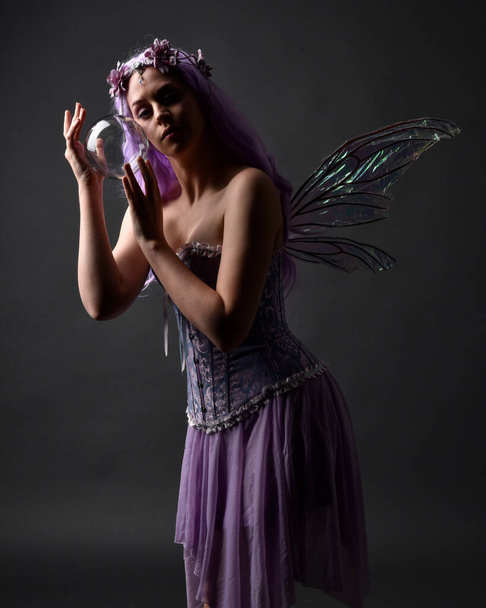 Κοντινό πορτραίτο ενός μοβ κοριτσιού με φανταστικό κορσέ φόρεμα με φτερά νεράιδας και στεφάνι λουλουδιών, που κάνει μάγια. Τοποθέτηση σε σκοτεινό φόντο στούντιο με σκιασμένο φωτισμό backlight. - Φωτογραφία, εικόνα