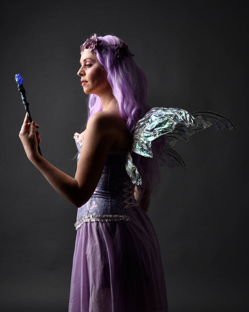 Close up portret fioletowe włosy dziewczyny w fantazji gorset sukienka z bajki skrzydła i korony kwiatowej, rzucając zaklęcie. Pozowanie na ciemnym tle studio z zacienionym oświetleniem podświetlanym. - Zdjęcie, obraz