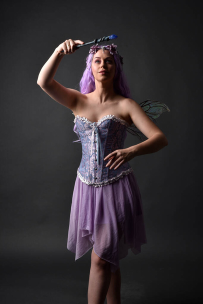 Gros plan portrait d'une fille aux cheveux violets portant une robe fantaisie corset avec des ailes de fées et une couronne de fleurs, jetant un sort. Poser sur un fond de studio sombre avec un éclairage rétroéclairé ombragé. - Photo, image