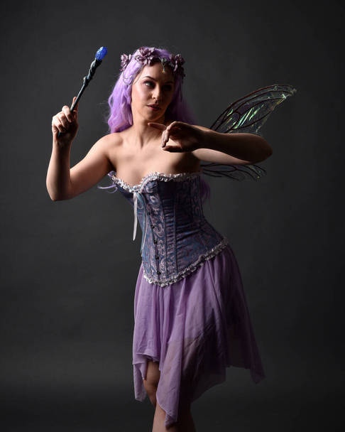 Close up portret fioletowe włosy dziewczyny w fantazji gorset sukienka z bajki skrzydła i korony kwiatowej, rzucając zaklęcie. Pozowanie na ciemnym tle studio z zacienionym oświetleniem podświetlanym. - Zdjęcie, obraz