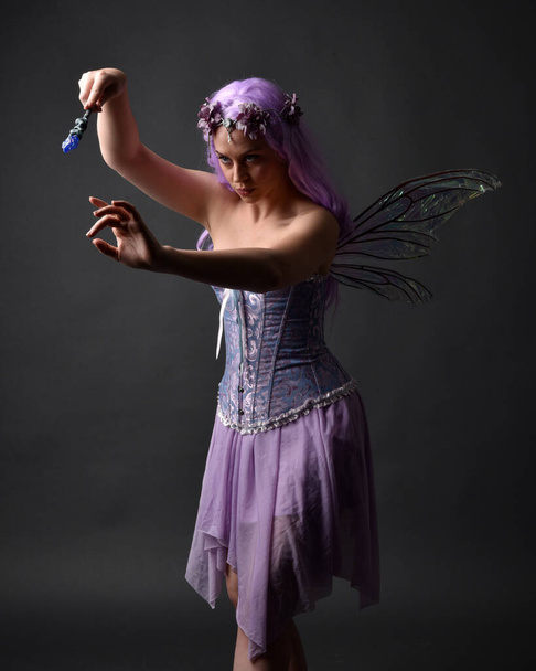 Close up portrait of a purple haired  girl wearing fantasy corset dress with fairy wings and flower crown, casting a spell.  Posing against a dark studio background with shadowed backlit lighting. - Fotografie, Obrázek