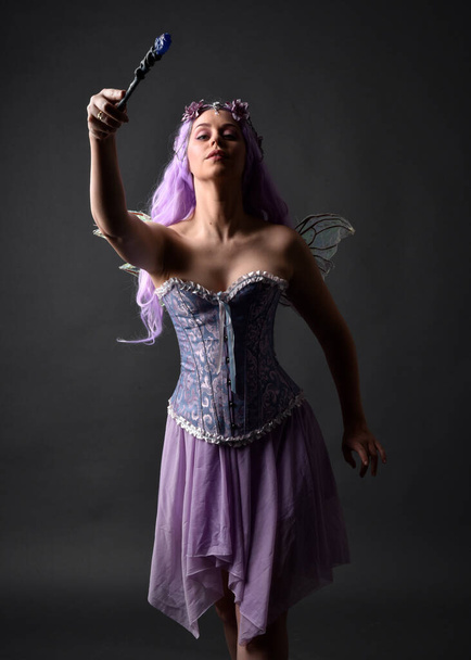 Close up portrait of a purple haired  girl wearing fantasy corset dress with fairy wings and flower crown, casting a spell.  Posing against a dark studio background with shadowed backlit lighting. - Photo, Image