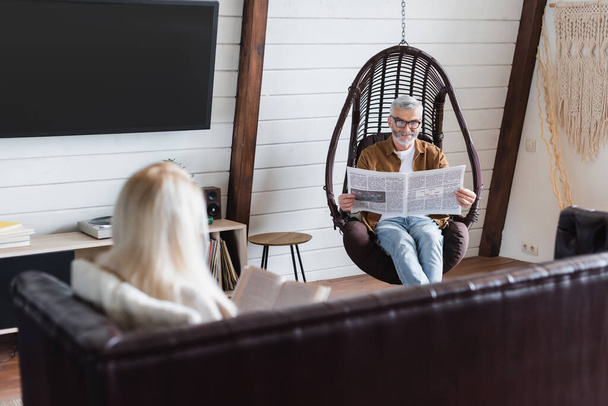 Старший чоловік читає новини у висячому кріслі біля розмитої дружини з книгою у вітальні
  - Фото, зображення