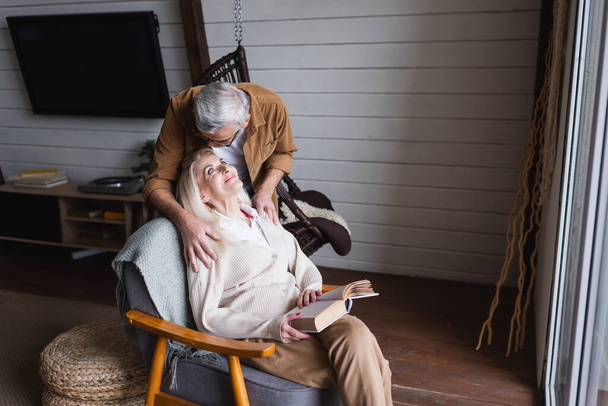 Elderly man hugging smiling wife with book in armchair at home  - Fotó, kép