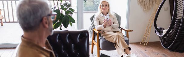 Alegre anciana usando teléfono inteligente en sillón cerca de marido borroso en la sala de estar, pancarta  - Foto, Imagen