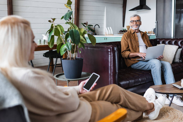 Sonriente anciano usando portátil cerca de esposa borrosa con teléfono celular en la sala de estar  - Foto, Imagen