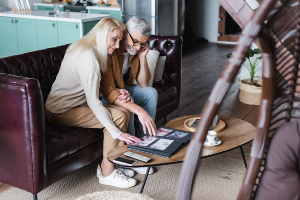 Senior couple looking at photos in album near smartphone on coffee table  - Fotoğraf, Görsel