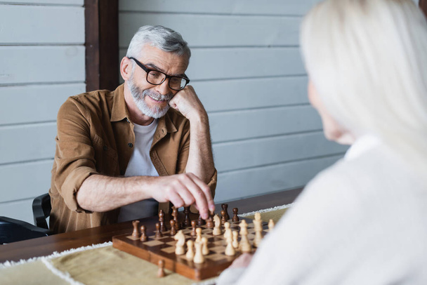 Χαμογελαστός ηλικιωμένος που παίζει σκάκι με τη γυναίκα του σε θολή πρόσοψη  - Φωτογραφία, εικόνα